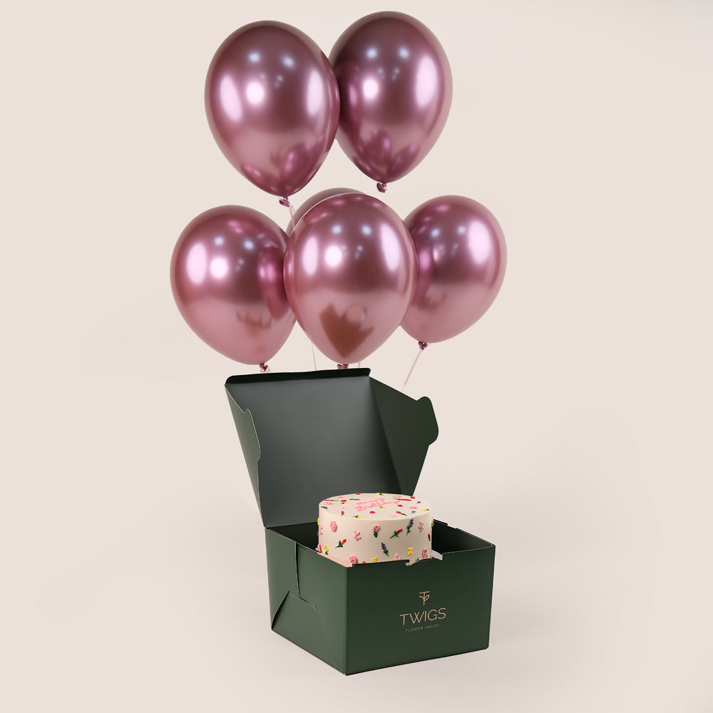 A 2kg floral birthday cake with intricate icing details, presented in a green box, accompanied by six metallic pink balloons.