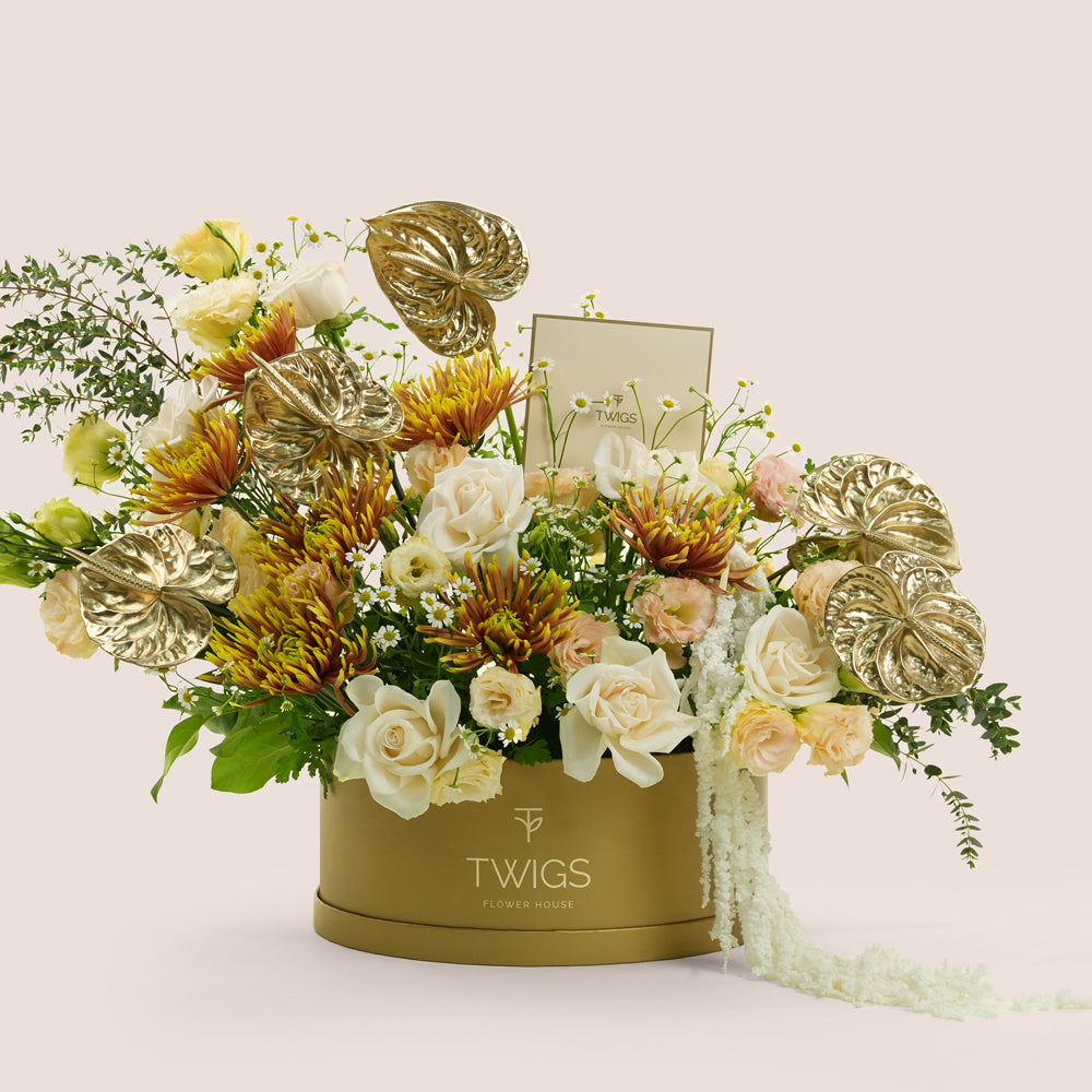 Aurelia, a luxury flower arrangement of white garden roses, saffina chrysanthemums, lisianthus, and gold painted anthurium leaves in a round TWIGS Gold box, with matching gift card.
