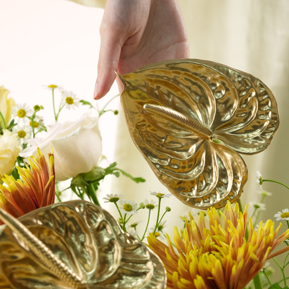 A hand holds a metallic gold anthurium-shaped decorative piece above a matching gold piece resting on a bouquet of white roses, yellow chrysanthemums, and small white daisies.