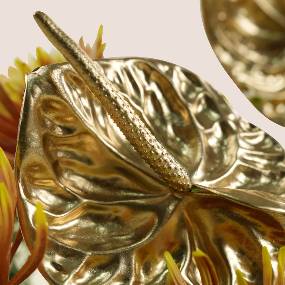 A close-up of a metallic gold anthurium-style decorative leaf with a textured central spike, shown among blurred yellow and orange flowers against a soft light beige background.