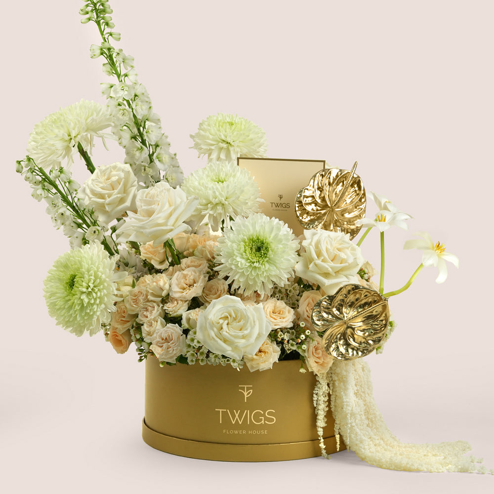Lumina, a luxury floral arrangement of white garden roses, deco chrysanthemums, salinero spray roses, delphiniums, tulips, and gold painted anthurium leaves in a round TWIGS Gold box, with matching gift card.