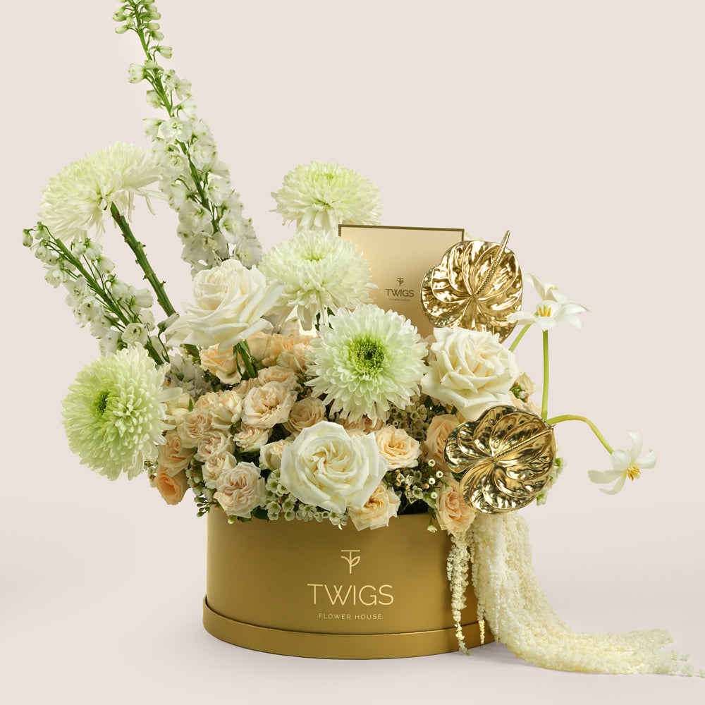 Lumina, a luxury floral arrangement of white garden roses, deco chrysanthemums, salinero spray roses, delphiniums, tulips, and gold painted anthurium leaves in a round TWIGS Gold box, with matching gift card.