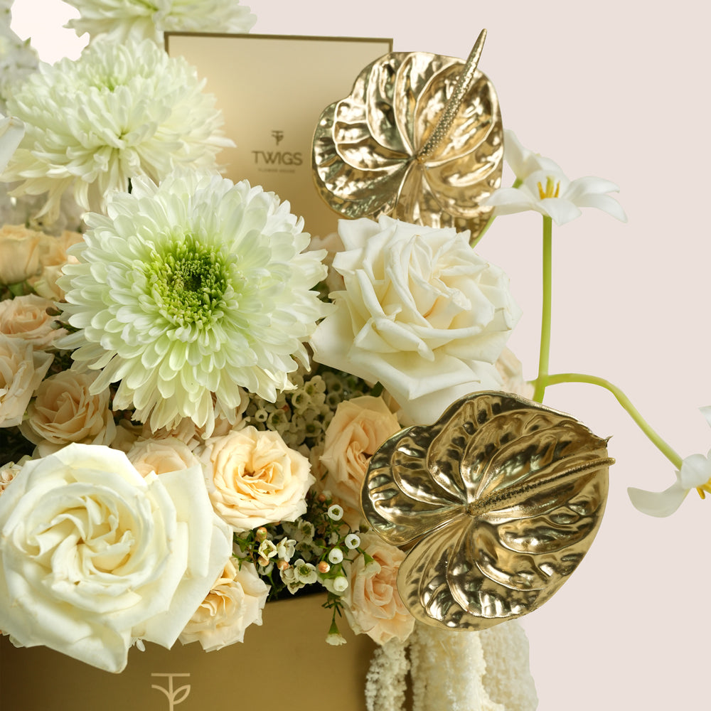 A close-up of a floral arrangement in a gold box with white and cream flowers, including chrysanthemums, roses, and white lilies, accented by two metallic gold leaf-shaped ornaments. The background is a soft light pink.
