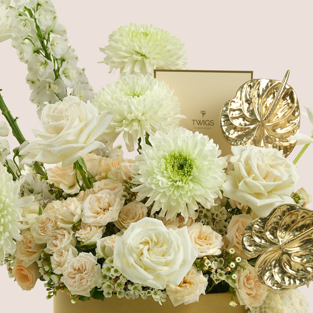 A luxury floral arrangement featuring white and cream roses, chrysanthemums, and delphiniums in a gold box, accented by two gold anthuriums, with a gold TWIGS box visible behind the flowers.