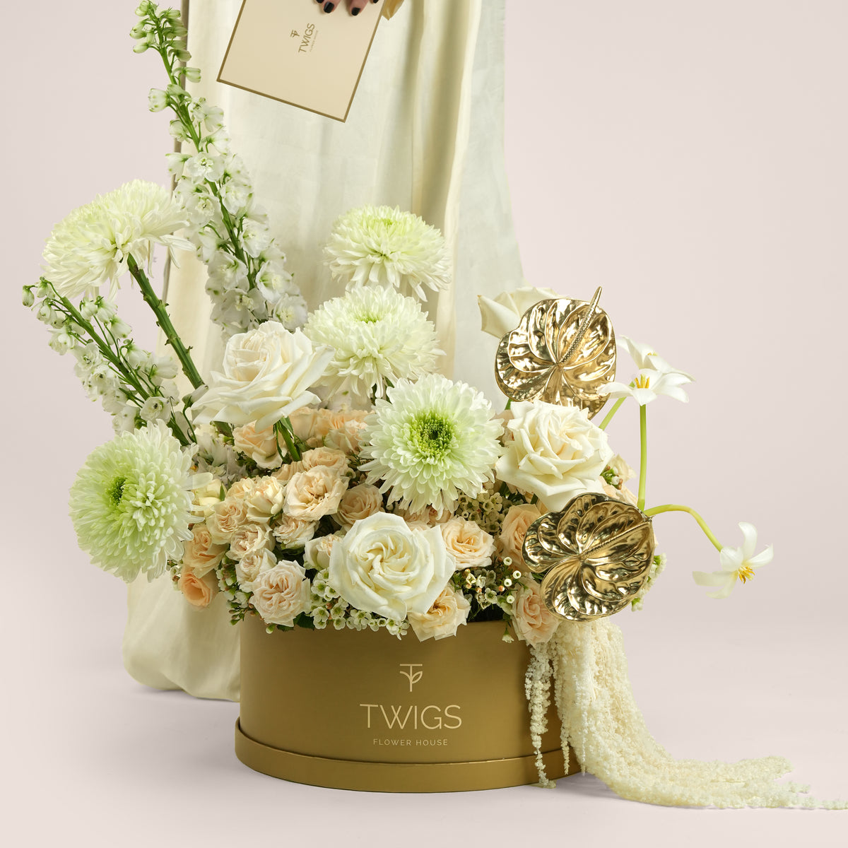Lumina, a luxury floral arrangement of white garden roses, deco chrysanthemums, salinero spray roses, delphiniums, tulips, and gold painted anthurium leaves in a round TWIGS Gold box, with matching gift card.