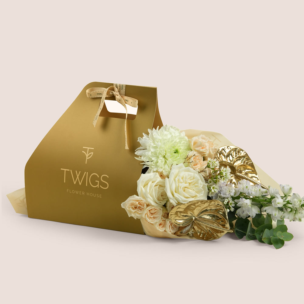 Lumina, a TWIGS Gold carry bag with white roses, cream spray roses, chrysanthemums, delphiniums, and a metallic gold anthurium leaf accent, on a light beige background.