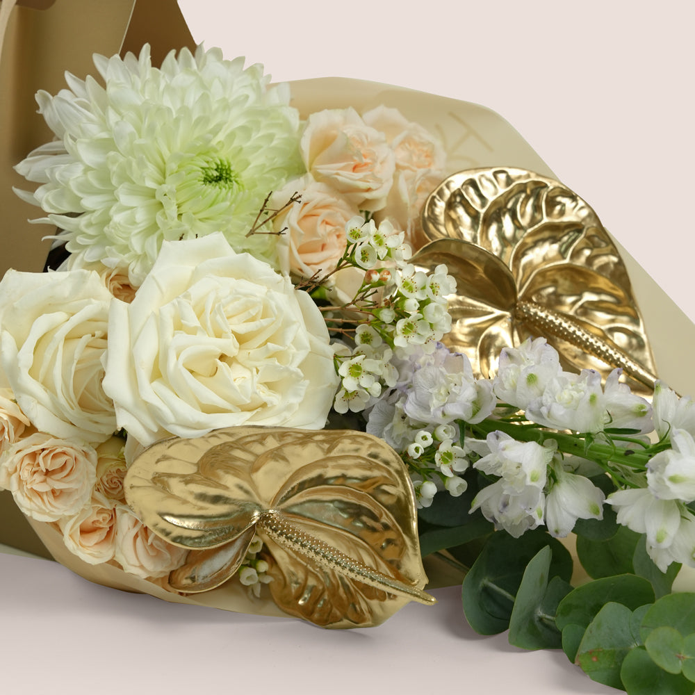 A close-up of a floral arrangement with white and blush peach roses, white snapdragons, green eucalyptus leaves, and two metallic gold decorative leaf ornaments wrapped in beige paper.