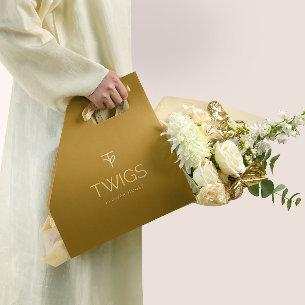Lumina, a TWIGS Gold carry bag with white roses, cream spray roses, chrysanthemums, delphiniums, and a metallic gold anthurium leaf accent, on a light beige background.