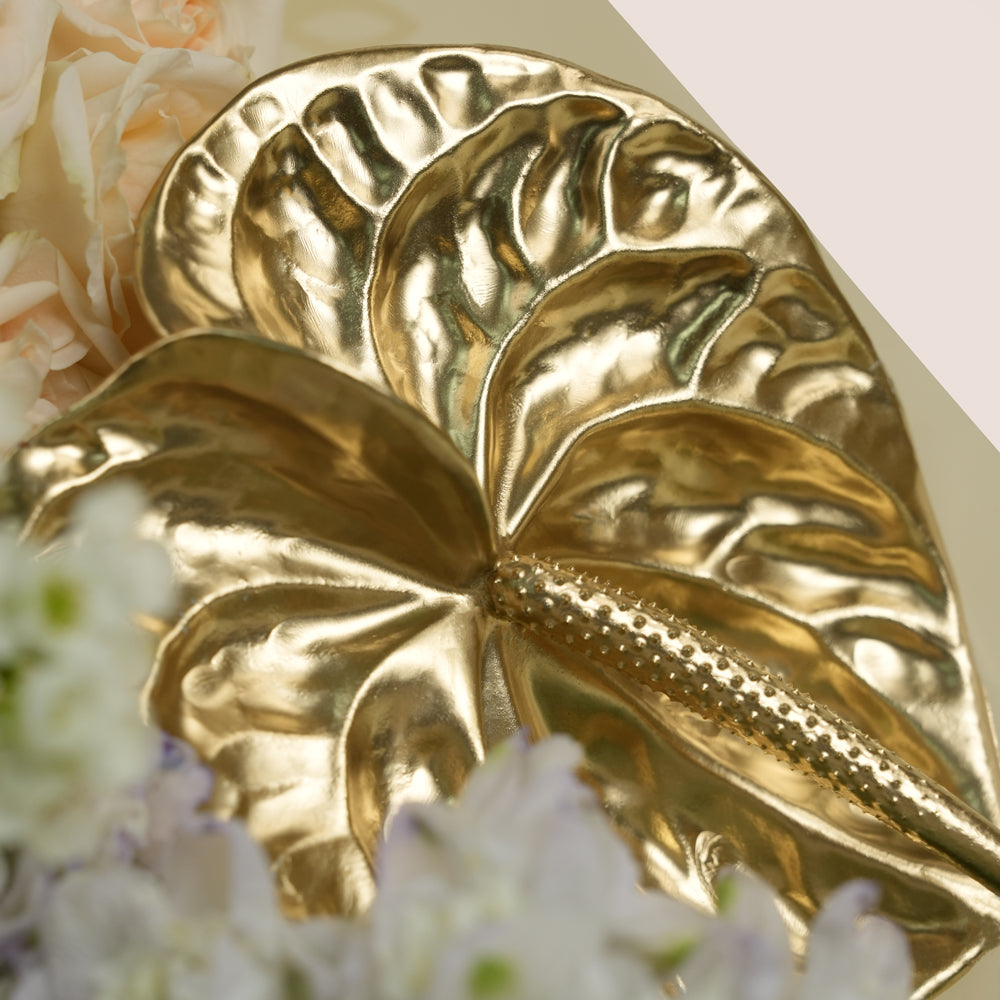 A metallic gold decorative leaf-shaped object with a textured, glossy finish, styled among soft white and pale pink flowers.