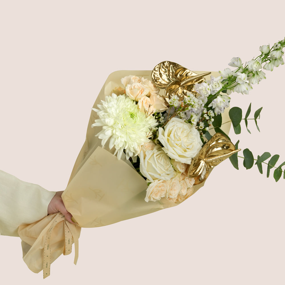Lumina, a TWIGS Gold carry bag with white roses, cream spray roses, chrysanthemums, delphiniums, and a metallic gold anthurium leaf accent, on a light beige background.