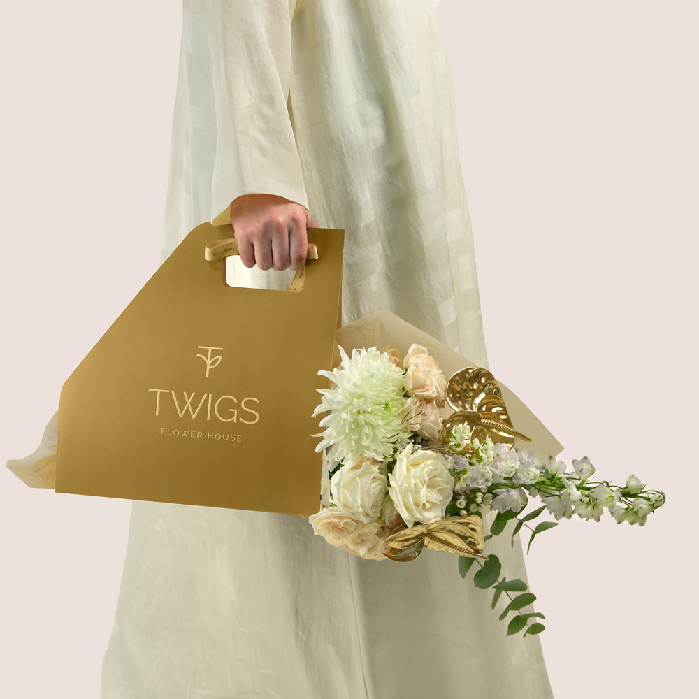 Lumina, a TWIGS Gold carry bag with white roses, cream spray roses, chrysanthemums, delphiniums, and a metallic gold anthurium leaf accent, on a light beige background.