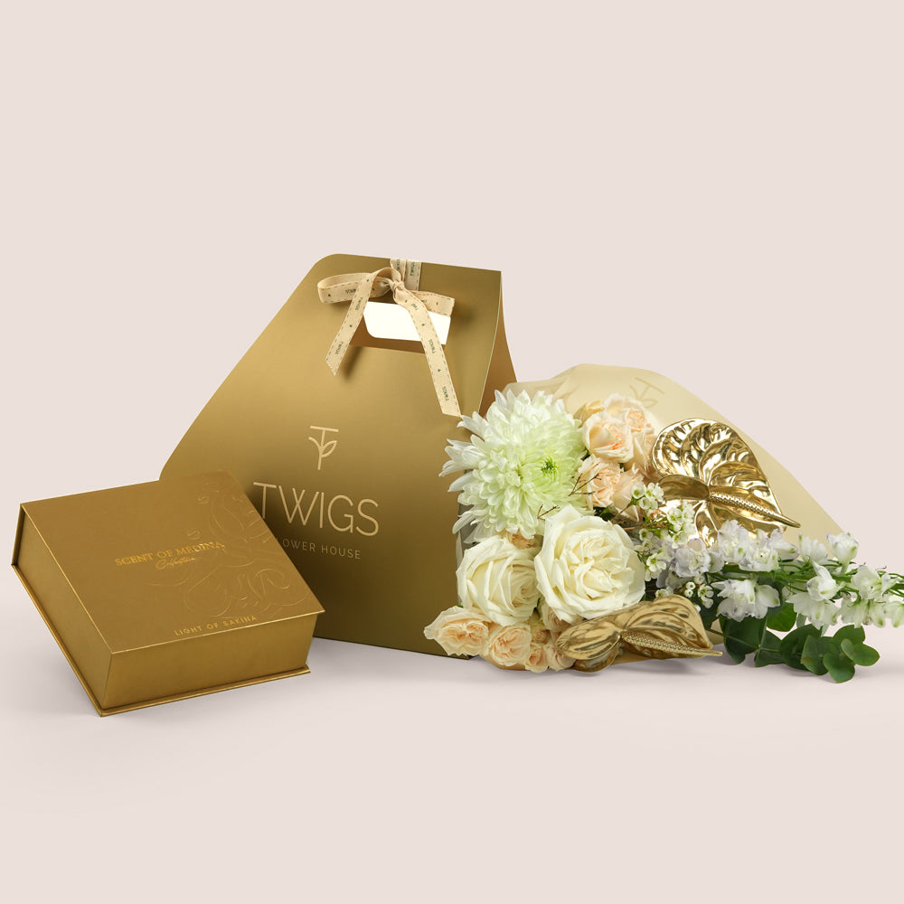 Lumina, a TWIGS Gold carry bag with white garden roses, cream spray roses, chrysanthemums, delphiniums, and a gold anthurium leaf accent, paired with the Contemplation Gift Set by Light of Sakina.