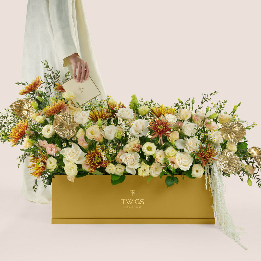 Aurelia, a luxury floral arrangement of white roses, chrysanthemums, lisianthus, and gold painted anthurium leaves in a rectangular TWIGS Gold garden box, with matching gift card.