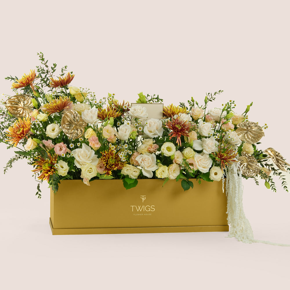 Aurelia, a luxury floral arrangement of white roses, chrysanthemums, lisianthus, and gold painted anthurium leaves in a rectangular TWIGS Gold garden box, with matching gift card.