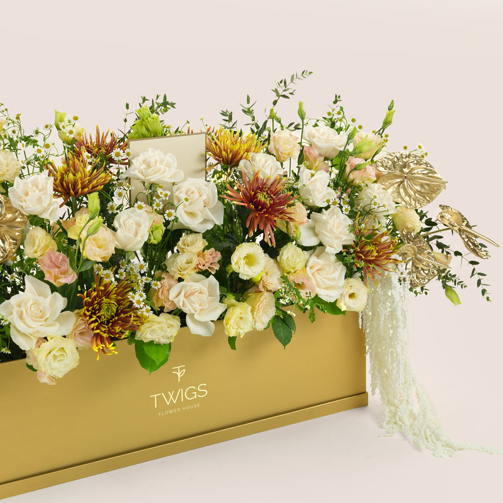 Aurelia, a luxury floral arrangement of white roses, chrysanthemums, lisianthus, and gold painted anthurium leaves in a rectangular TWIGS Gold garden box, with matching gift card.