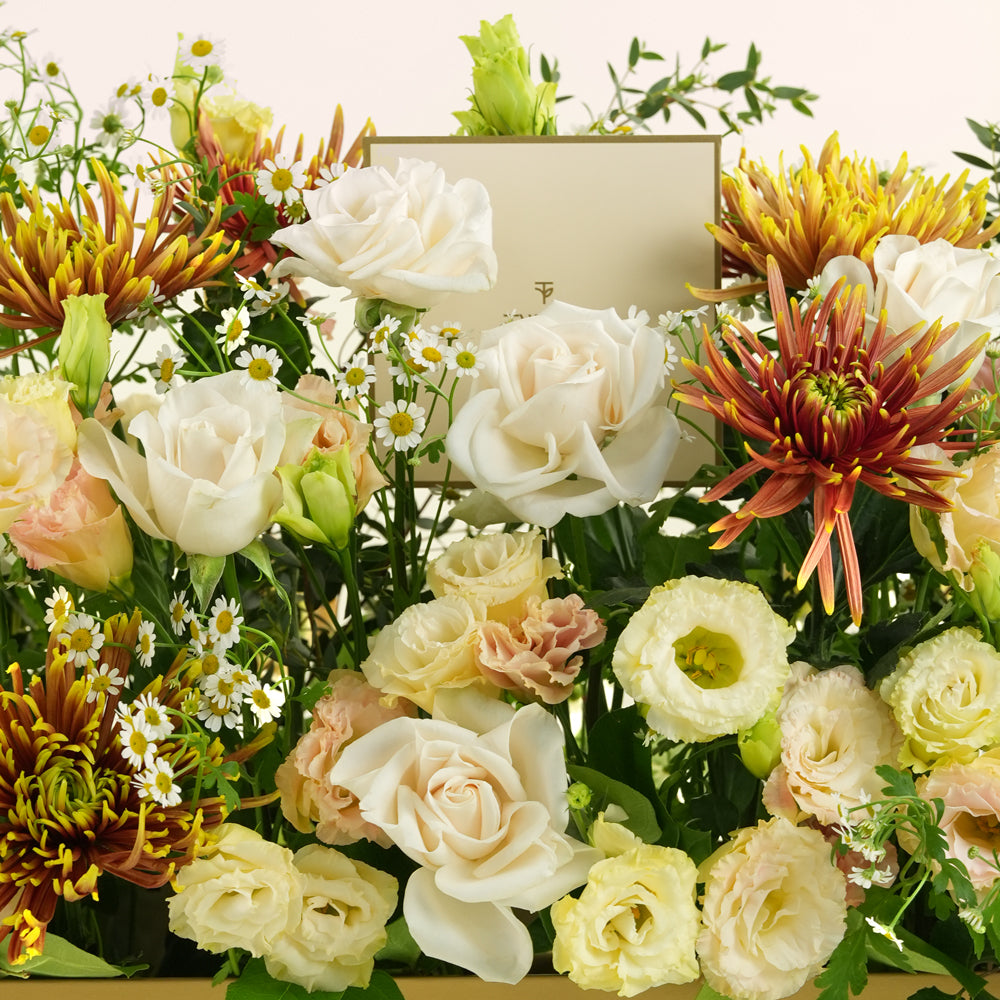 A dense floral arrangement featuring cream and white roses, pale peach lisianthus, orange and yellow chrysanthemums, and small white daisy-like flowers with green foliage, set against a light neutral background.