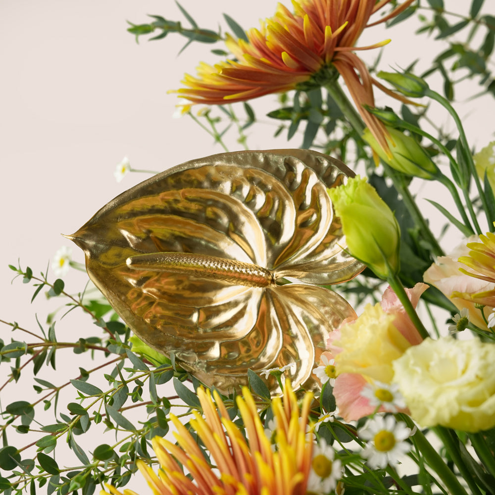 A decorative gold leaf-shaped wall hanging or ornament displayed among a colorful floral arrangement with orange, yellow, pink, white, and green flowers against a light beige background.