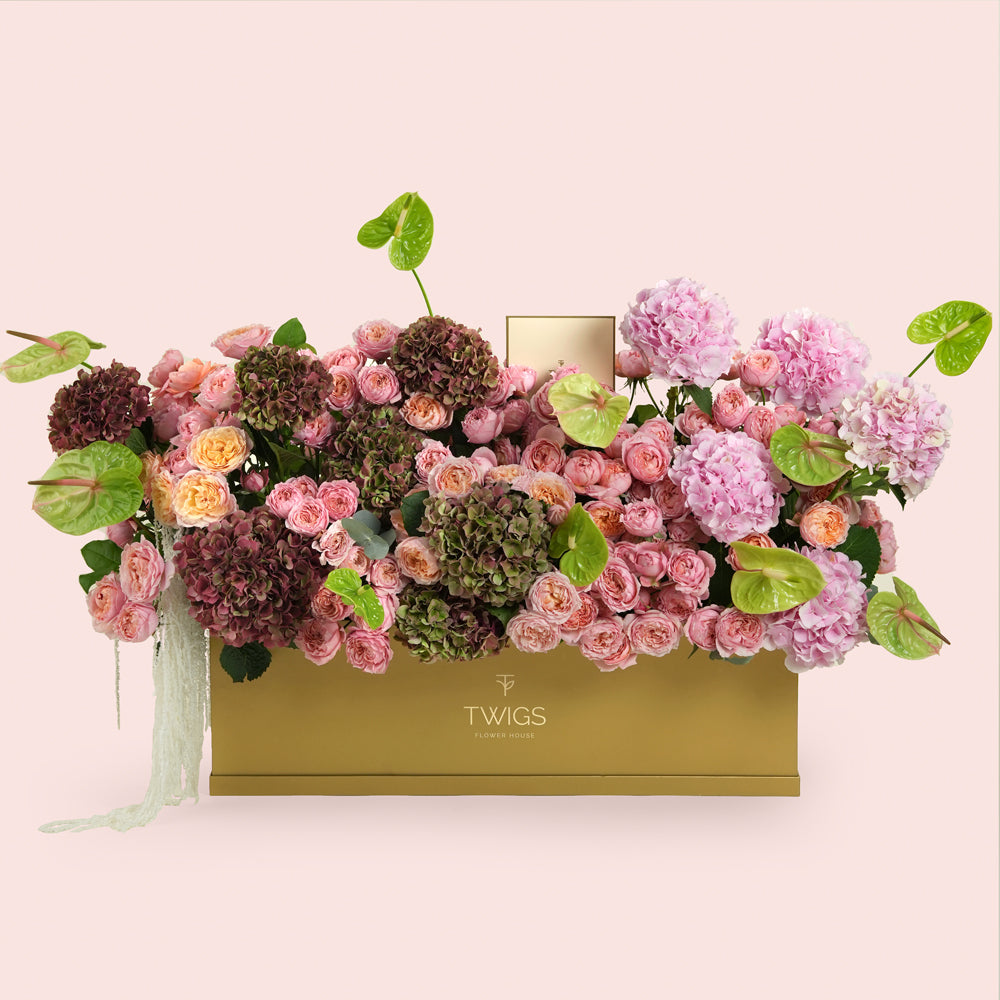 Isadora, a luxury floral arrangement of pink and peach garden roses, pink and classic hydrangeas, and green anthurium leaves in a rectangular TWIGS Gold garden box.