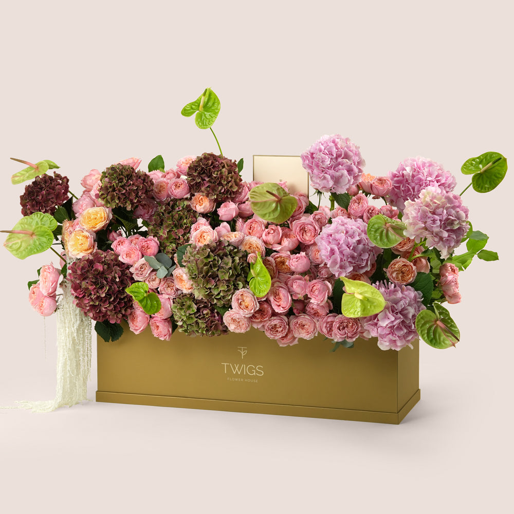 Isadora, a luxury floral arrangement of pink and peach garden roses, pink and classic hydrangeas, and green anthurium leaves in a rectangular TWIGS Gold garden box.