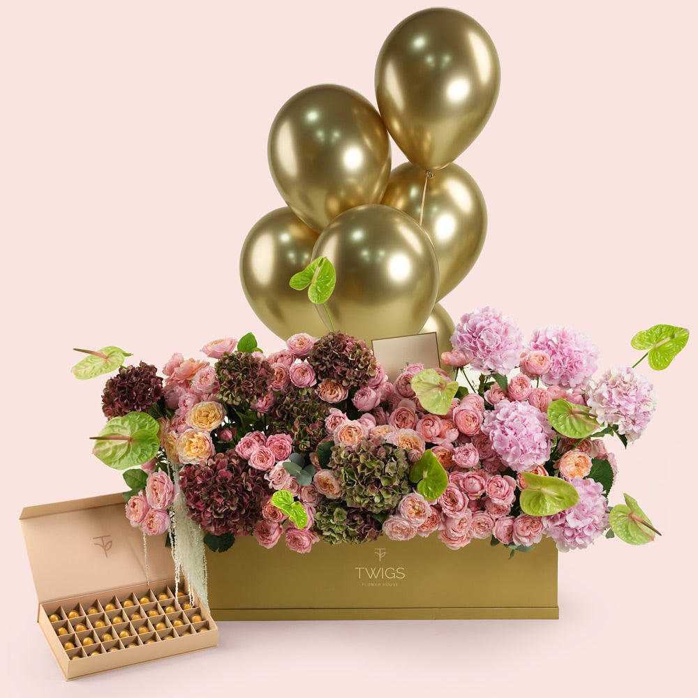 Isadora Royale, a luxury TWIGS Gold Garden box arrangement of pink and burgundy hydrangeas, garden roses, and green anthurium leaves, paired with a 32 piece premium bonbon box and six gold chrome balloons.