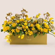 Solara, a vibrant yellow floral arrangement of calla lilies, roses, chrysanthemums, kangaroo paw, and gold painted anthurium leaves in a rectangular TWIGS Gold garden box.