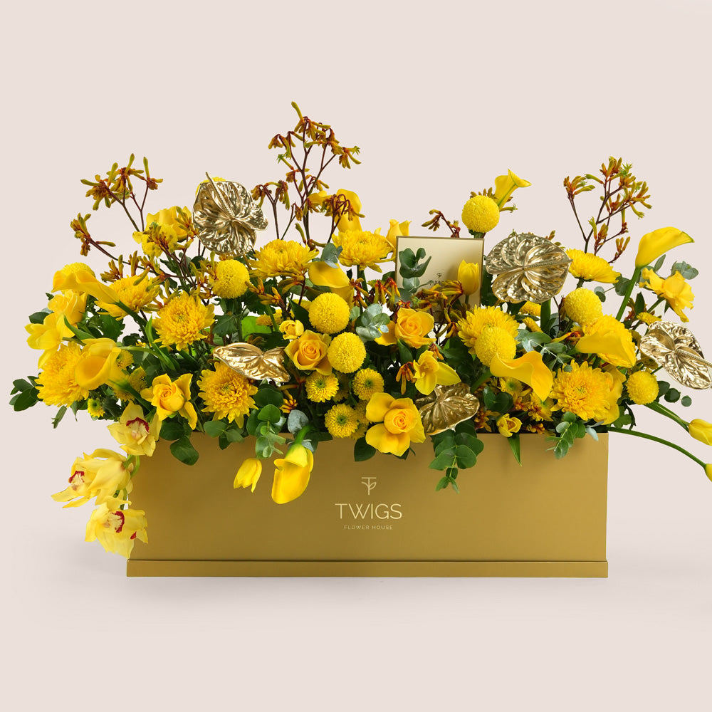 Solara, a vibrant yellow floral arrangement of calla lilies, roses, chrysanthemums, kangaroo paw, and gold painted anthurium leaves in a rectangular TWIGS Gold garden box.