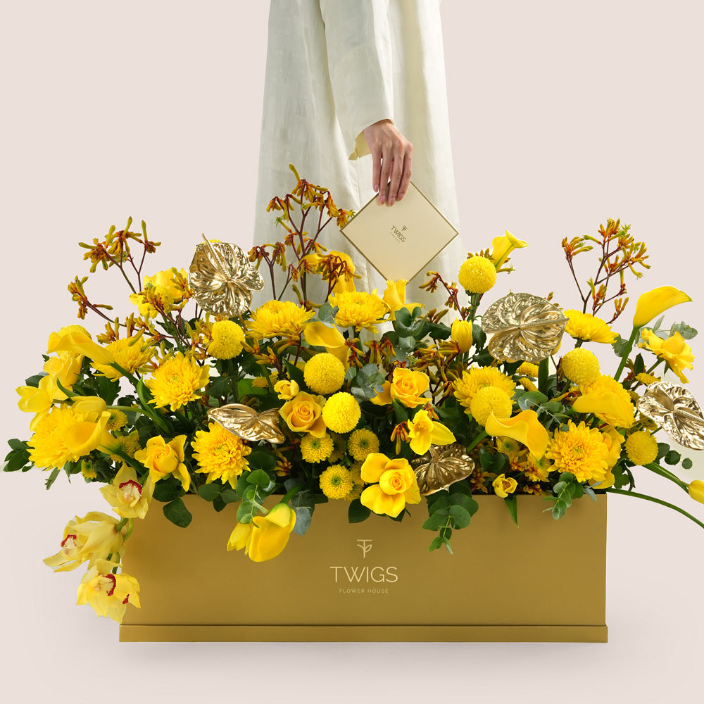Solara, a vibrant yellow floral arrangement of calla lilies, roses, chrysanthemums, kangaroo paw, and gold painted anthurium leaves in a rectangular TWIGS Gold garden box.