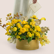Solara, a vibrant yellow floral arrangement of calla lilies, chrysanthemums, tulips, roses, cymbidium orchids, kangaroo paw, and gold painted anthurium leaves in a round TWIGS Gold flower box.