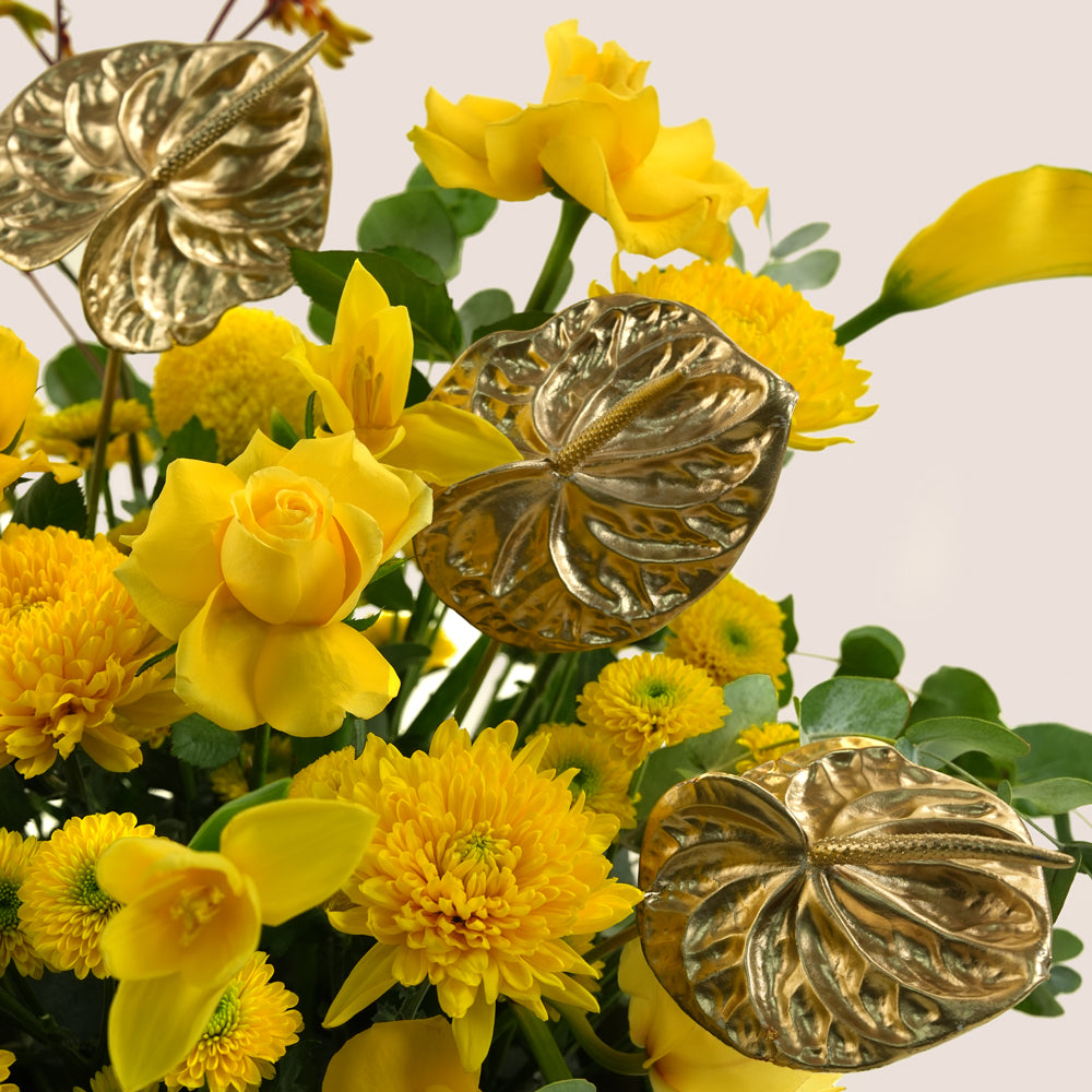 A close-up of a yellow floral arrangement featuring real yellow roses, mums, calla lilies, and green foliage, with three large metallic gold anthurium-style decorative leaves layered over the flowers against a light neutral background.