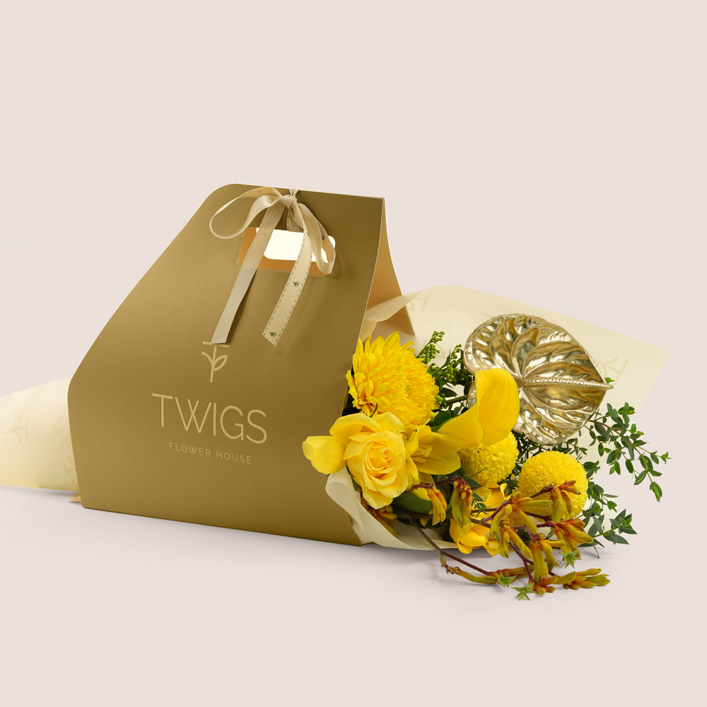 Solara, a TWIGS Gold carry bag with yellow roses, chrysanthemums, kangaroo paw, and a metallic gold anthurium leaf accent, on a light beige background.