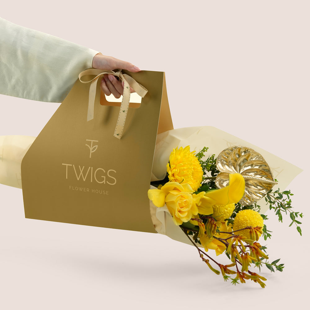 Solara, a TWIGS Gold carry bag with yellow roses, chrysanthemums, kangaroo paw, and a metallic gold anthurium leaf accent, on a light beige background.