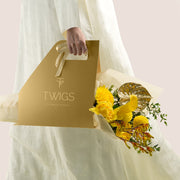 Solara, a TWIGS Gold carry bag with yellow roses, chrysanthemums, kangaroo paw, and a metallic gold anthurium leaf accent, on a light beige background.