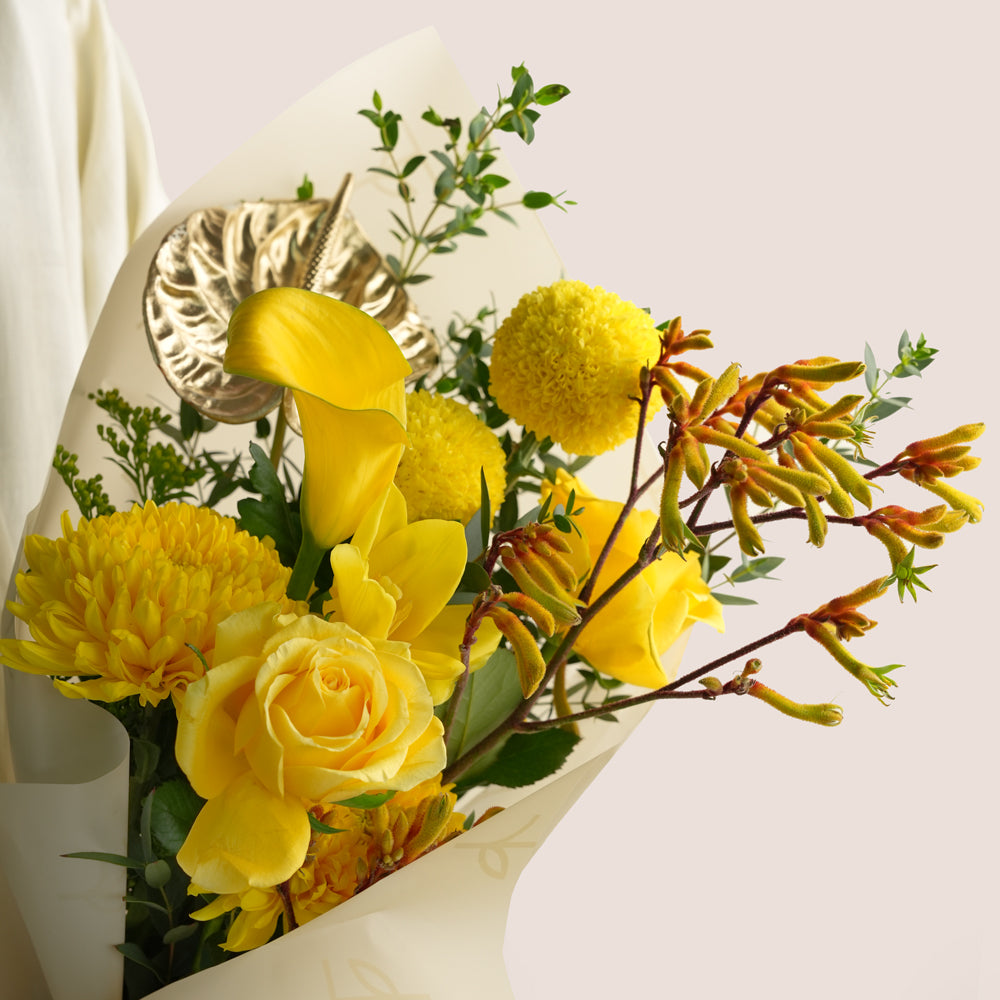 A close-up of a yellow flower bouquet wrapped in cream paper, featuring yellow roses, calla lilies, chrysanthemums, and orange blossoms with green foliage, set against a light beige background.
