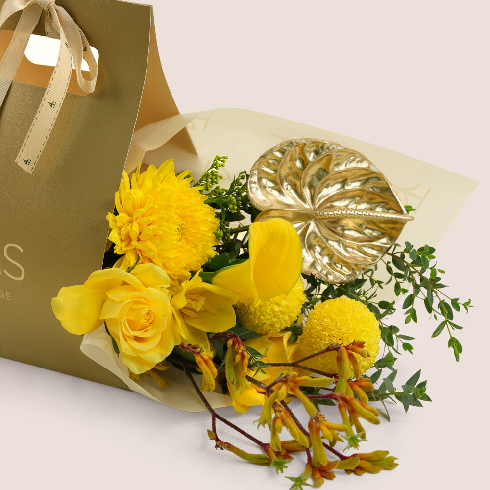 Solara, a TWIGS Gold carry bag with yellow roses, chrysanthemums, kangaroo paw, and a metallic gold anthurium leaf accent, on a light beige background.