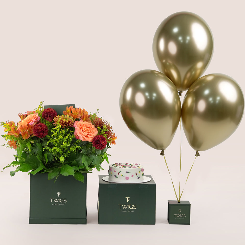 Amber Harvest square flower box with orange and burgundy flowers, paired with a 1kg birthday cake and three chrome gold balloons in TWIGS beige or green packaging, available for same day flower delivery in Dubai when you buy flowers online.