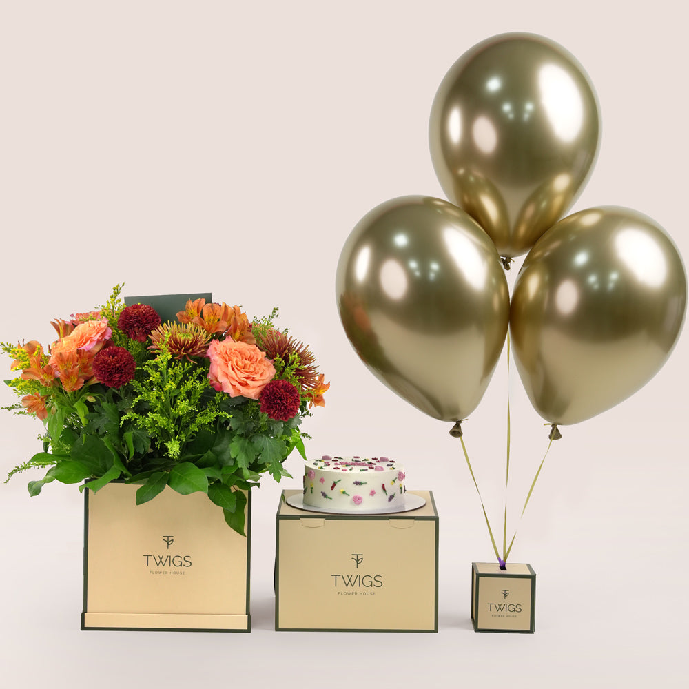 Amber Harvest square flower box with orange and burgundy flowers, paired with a 1kg birthday cake and three chrome gold balloons in TWIGS beige or green packaging, available for same day flower delivery in Dubai when you buy flowers online.