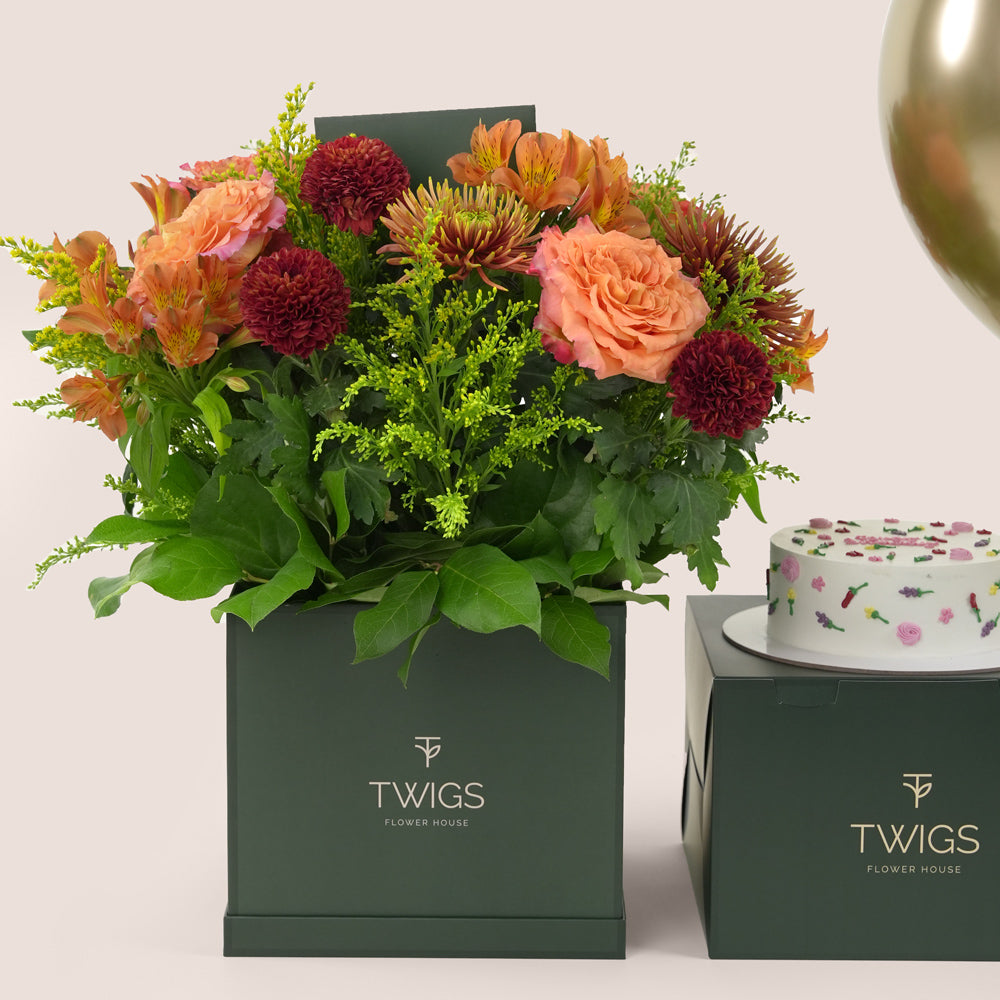 Amber Harvest square flower box with orange and burgundy flowers, paired with a 1kg birthday cake and three chrome gold balloons in TWIGS beige or green packaging, available for same day flower delivery in Dubai when you buy flowers online.