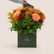 Orange Ecuador roses, alstroemerias, spider chrysanthemums, and burgundy pom pom mums arranged with greenery in a TWIGS beige or green square box, available for same day flower delivery in Dubai when you buy flowers online.