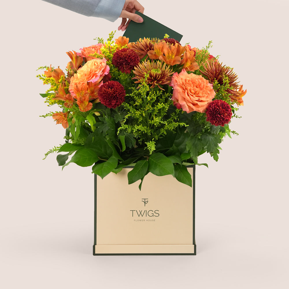 Orange Ecuador roses, alstroemerias, spider chrysanthemums, and burgundy pom pom mums arranged with greenery in a TWIGS beige or green square box, available for same day flower delivery in Dubai when you buy flowers online.
