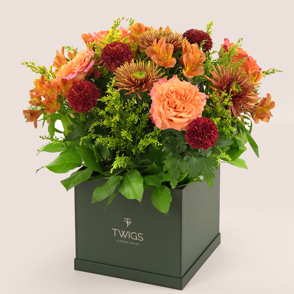 Orange Ecuador roses, alstroemerias, spider chrysanthemums, and burgundy pom pom mums arranged with greenery in a TWIGS beige or green square box, available for same day flower delivery in Dubai when you buy flowers online.