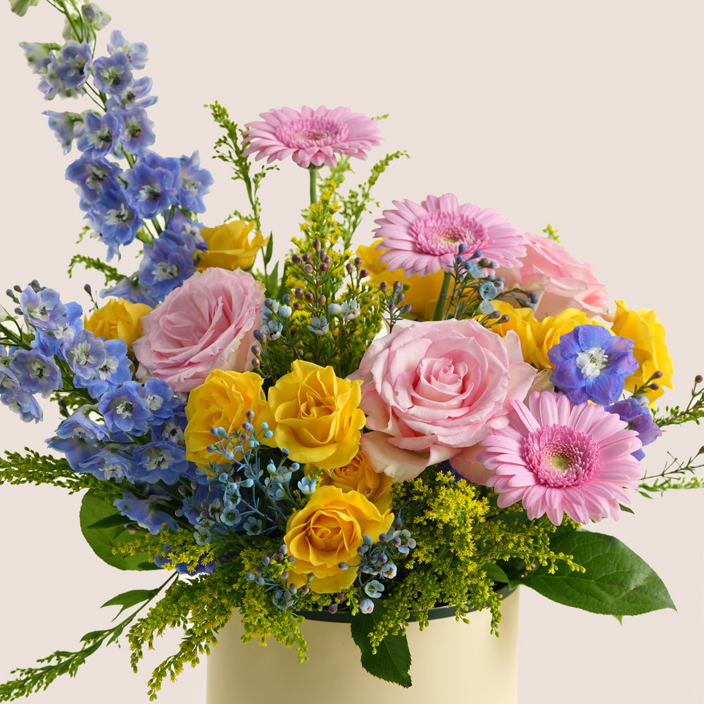 Pink and yellow rose arrangement with gerbera daisies and blue delphiniums in a beige TWIGS Flower House box, Sunday Bloom Deluxe