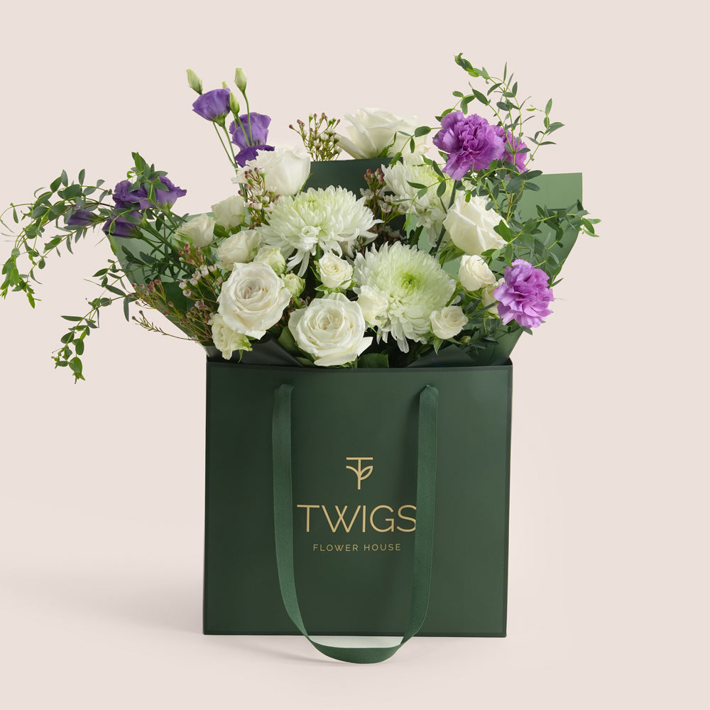 White rose and purple lisianthus bouquet in a green TWIGS Flower House signature bag on a beige background