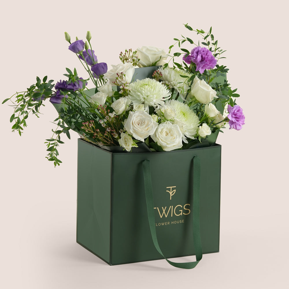 White rose and purple lisianthus bouquet in a green TWIGS Flower House signature bag on a beige background