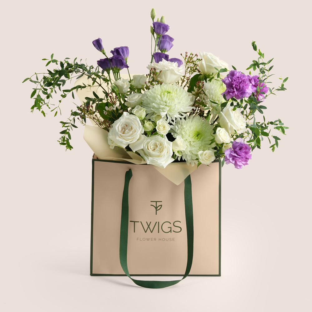 White rose and purple lisianthus bouquet in a green TWIGS Flower House signature bag on a beige background