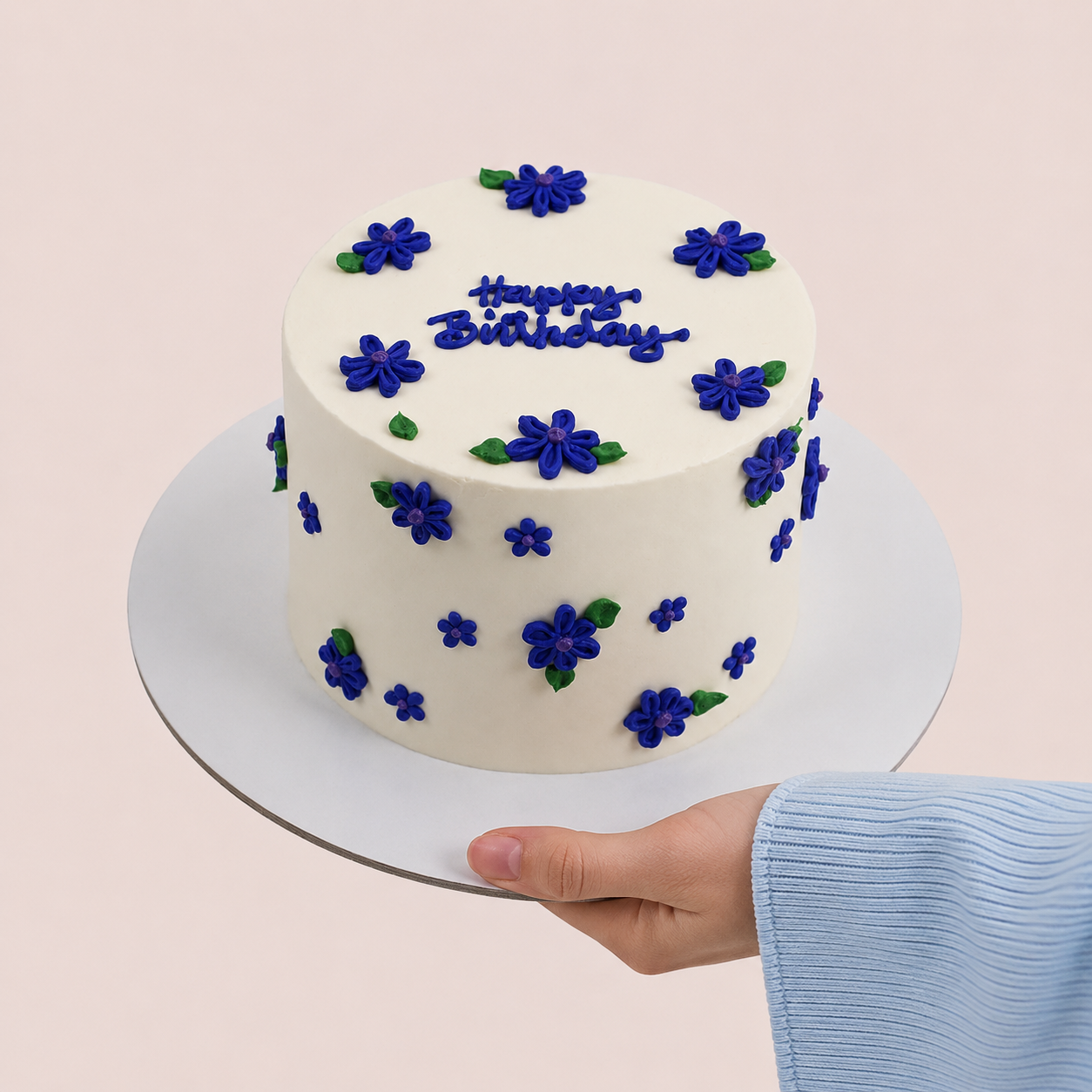 A round white frosted cake decorated with blue flower motifs and green leaves, topped with blue icing text reading 'Happy Birthday,' shown on a white cake board being held by a hand wearing a light blue ribbed sleeve against a pale pink background.
