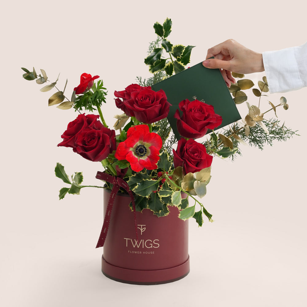 Bouquet of red roses and red poppies in a maroon container with 'TWIGS' branding, held by a hand.