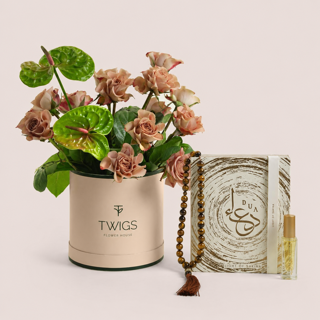 Café latte roses and green anthurium in a dark green TWIGS box beside a Light of Sakina Tiger Eye misbaha and perfume oil, TWIGS x Light of Sakina collaboration