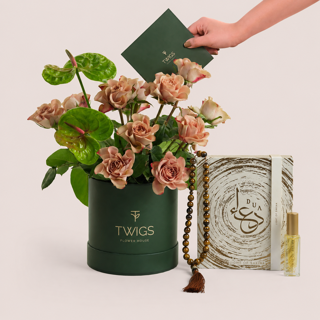Café latte roses and green anthurium in a dark green TWIGS box beside a Light of Sakina Tiger Eye misbaha and perfume oil, TWIGS x Light of Sakina collaboration
