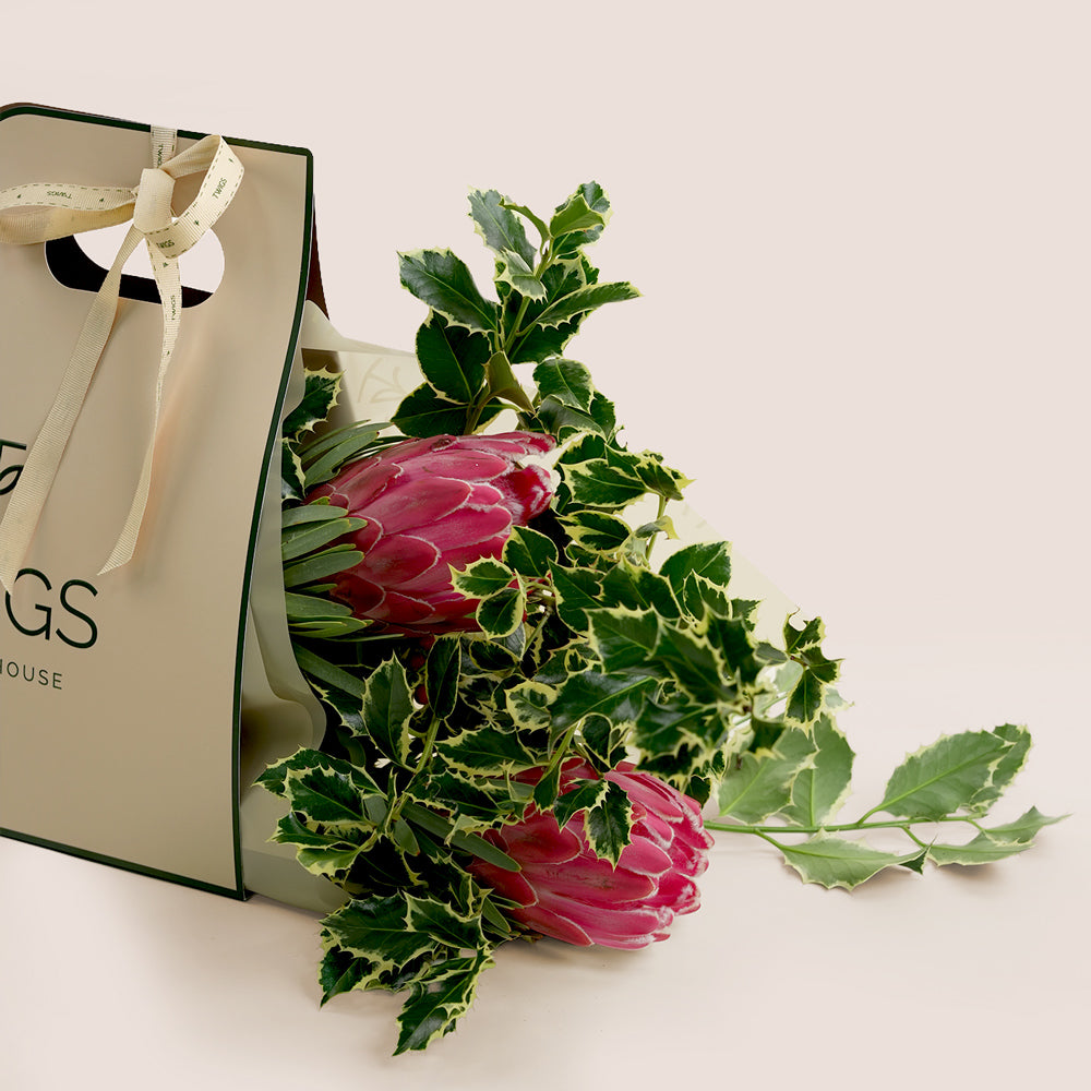 Floral arrangement with pink protea flowers and green leaves next to a branded gift bag on a light background