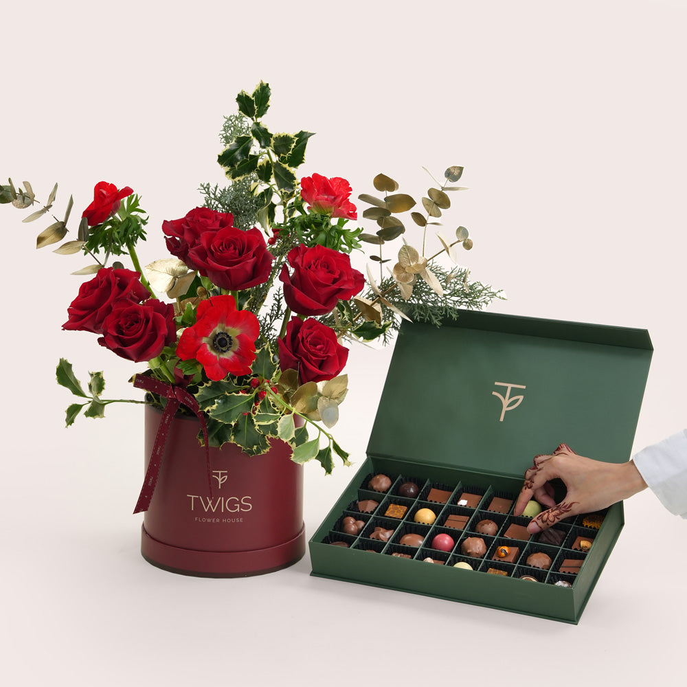 Bouquet of red roses and greenery in a maroon container with 'TWIGS' branding, next to an open green box of chocolates.